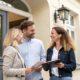 Pareja recibiendo asesoramiento en la compra de tu vivienda por parte de una agente inmobiliaria frente a la entrada de una casa, todos sonrientes mientras revisan documentos.